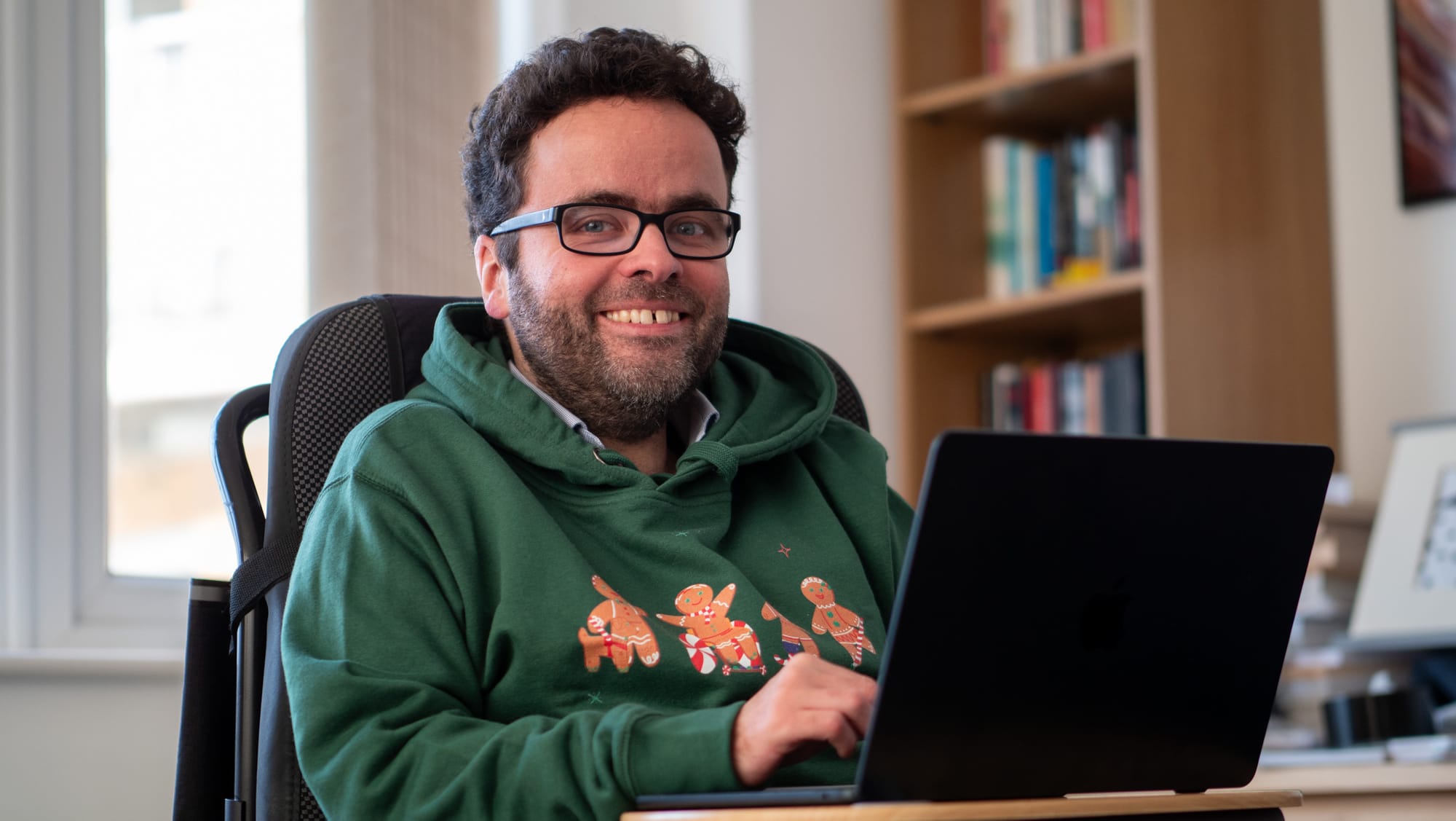 Photo of Peter, a white man nearly 40, sitting in wheelchair and engaging smile. He works on a laptop, and wears a green hoody with a disabled-gingerbreadman theme. He has short dark curly hair, glasses and a trimmed beard.