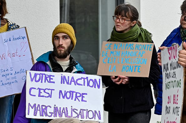 Two people in a group of protestors, holding signs in French. A white man with a beard sitting in a wheelchair holds a sign saying "Votre inaction c'est notre marginalisation". Next to him a white woman standing with a sign saying "Je n'ai pas honte de mon handicap, cést exclusion la honte."