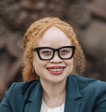 Profile picture of IK, a woman with albinism. She has an open smile, glasses with a dark frame, and light-red curly hair framing her face.